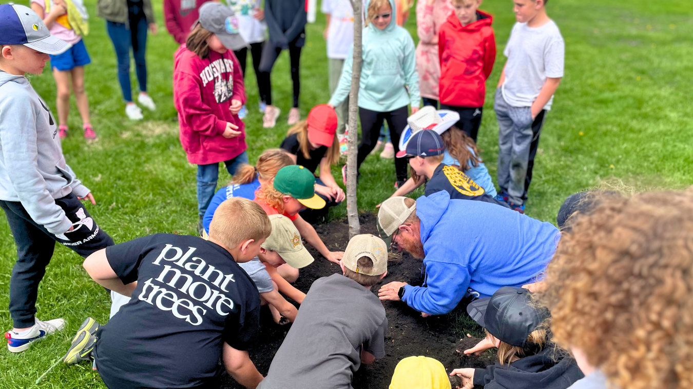 Children from a local school are seen helping to plant a tree for Arbor Day with one child wearing a shirt that says, plant more trees on the back.
